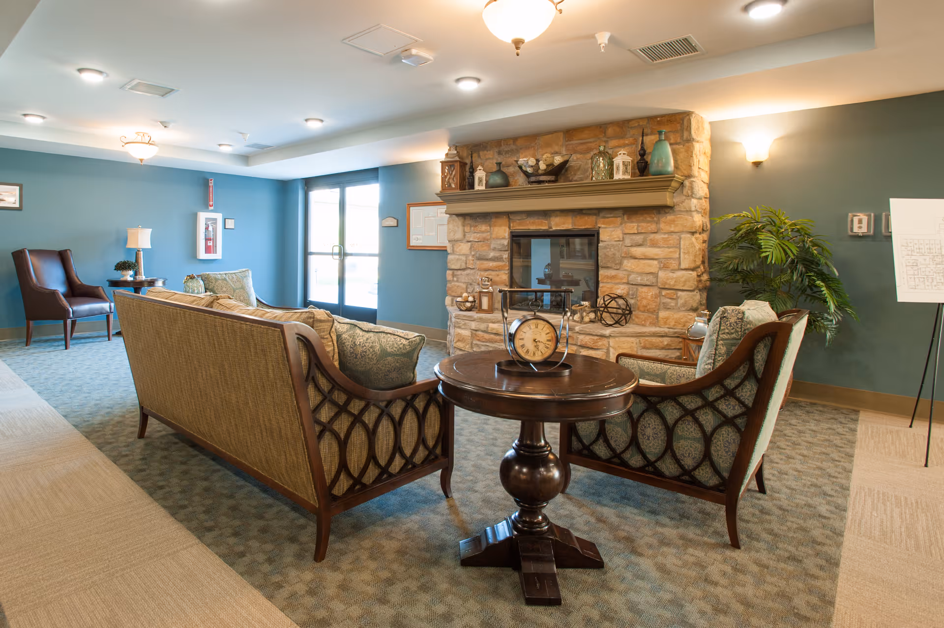 A cozy senior living facility common area with a stone fireplace, a wooden round table with a decorative clock, two upholstered armchairs, and a matching sofa. The walls are painted teal, and there is a large window letting in natural light. Decorative items and plants are placed around the room.