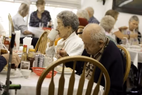 Elderly residents sitting at tables in a dining room, eating and drinking. The room has several tables with white tablecloths, condiments, and napkins. The residents appear engaged in their meals and conversations.