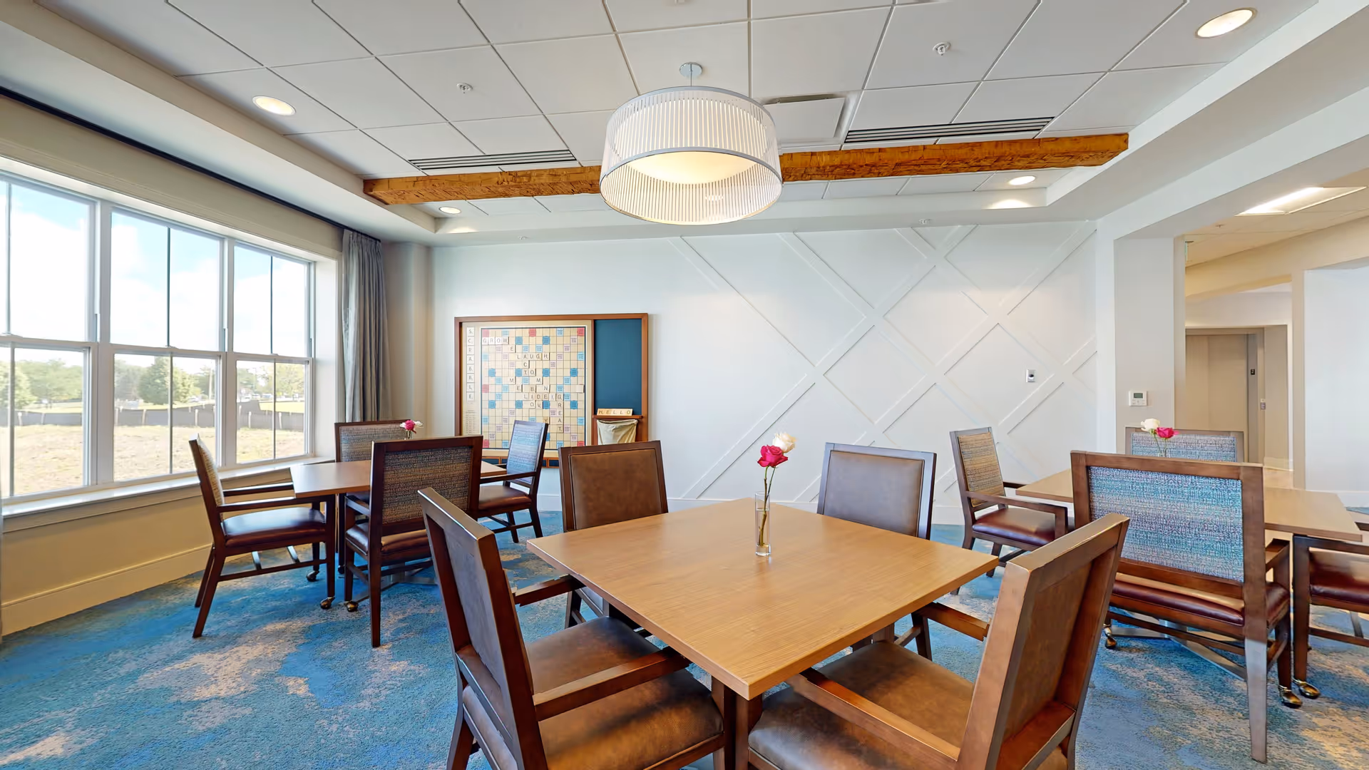 A bright and spacious dining room with several wooden tables and chairs arranged neatly. Each table has a small vase with a single flower. Large windows on the left side let in natural light, and a large Scrabble board is mounted on the far wall. The ceiling has recessed lighting and a modern hanging light fixture. The carpet is blue with a textured pattern.