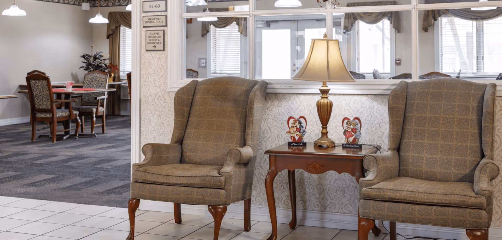 Two upholstered armchairs with wooden legs are positioned on either side of a wooden side table with a lamp and two decorative figurines. Behind the chairs is a window looking into a dining area with tables and chairs. The room has patterned wallpaper and tiled flooring near the chairs, with carpeted flooring in the dining area.