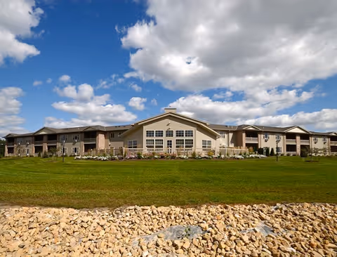 Wide front view of a low-rise memory care building set behind a green lawn and stone border under a blue, partly cloudy sky.