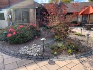 Outdoor patio area with paved walkways, garden beds containing shrubs and flowers, and outdoor seating with umbrellas. A building with brick walls and windows is visible in the background under a clear sky.