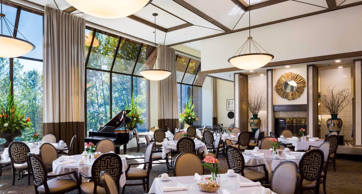Elegant dining room with round tables set for meals, upholstered chairs, large floor-to-ceiling windows, chandeliers, and a grand piano.