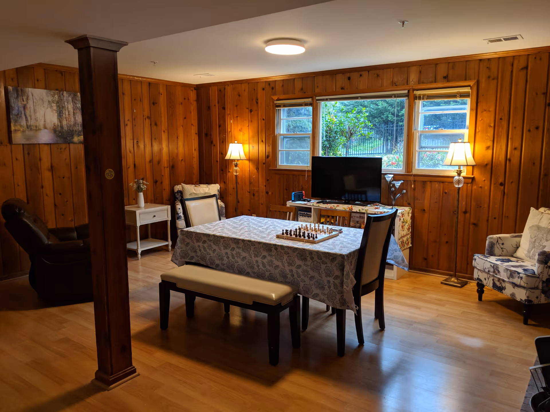 Wood-paneled common room with a table covered by a patterned tablecloth, a chessboard, chairs, TV and lamps by a window.