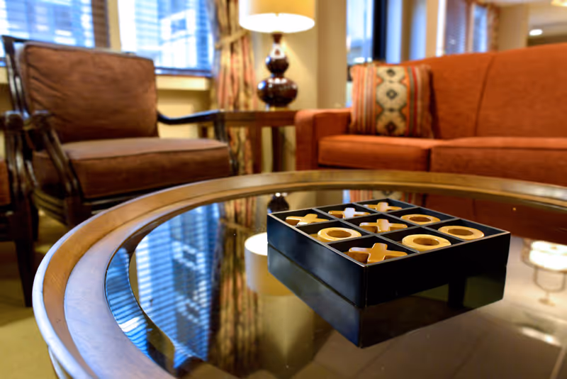 A cozy living room with a glass-top wooden coffee table in the foreground featuring a tic-tac-toe game set. In the background, there is a brown cushioned armchair, a red sofa with a patterned pillow, a wooden side table, and a lamp. Window blinds and curtains are visible behind the furniture.