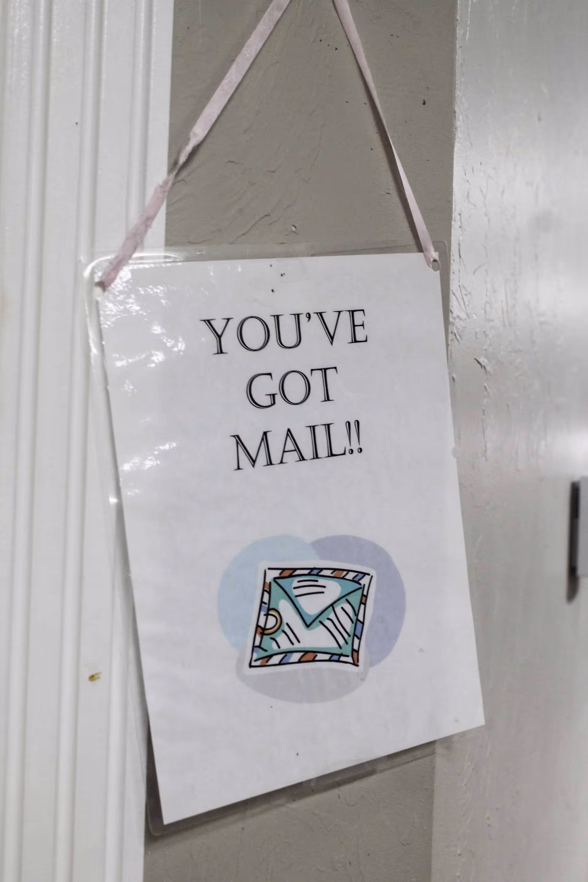 A laminated sign hanging on a wall with a pink ribbon that reads 'YOU'VE GOT MAIL!!' above a colorful illustration of an envelope.