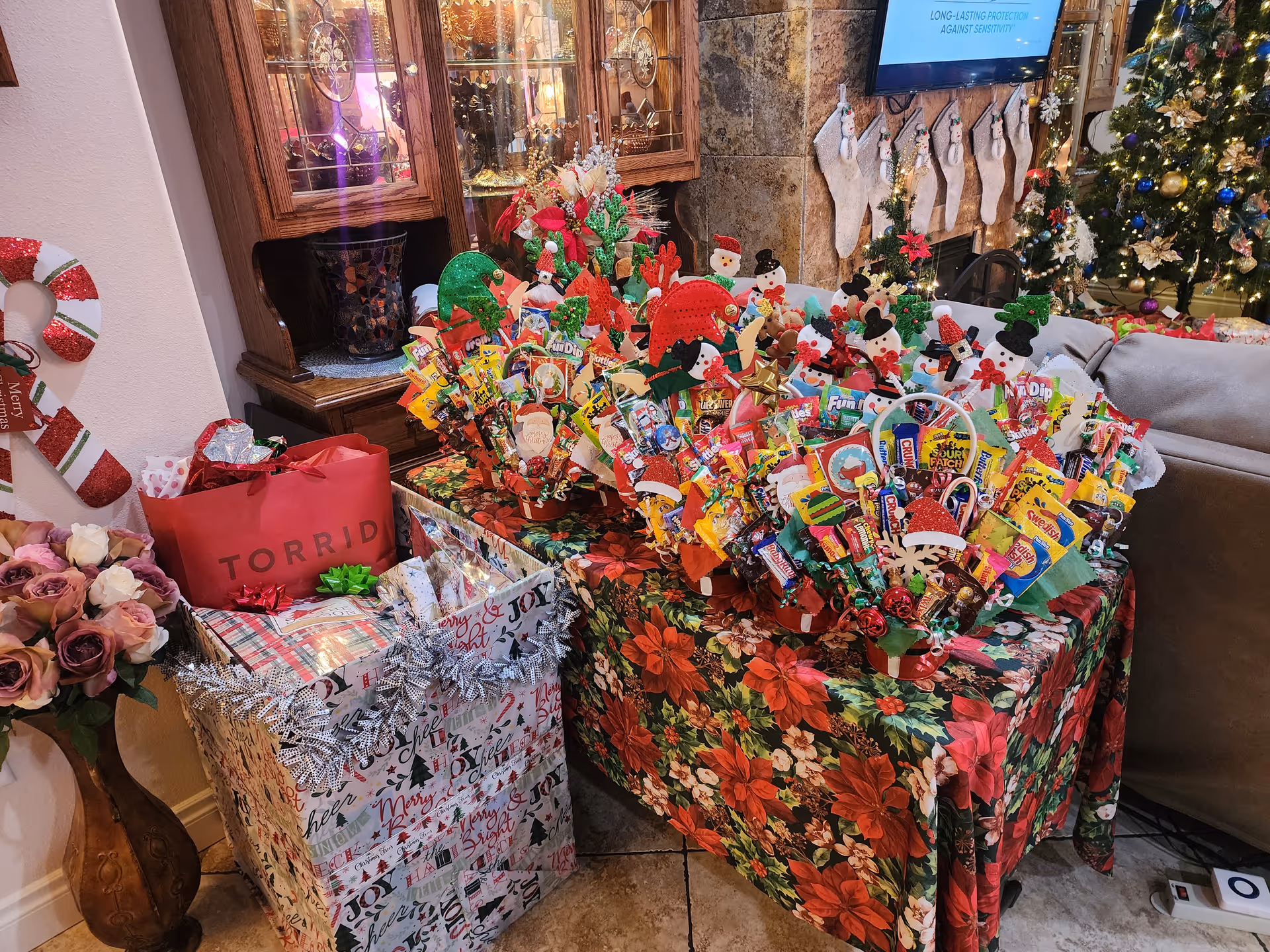 Festively decorated living room corner with wrapped gifts, candy bouquets, a Christmas tree, and stockings on a fireplace.