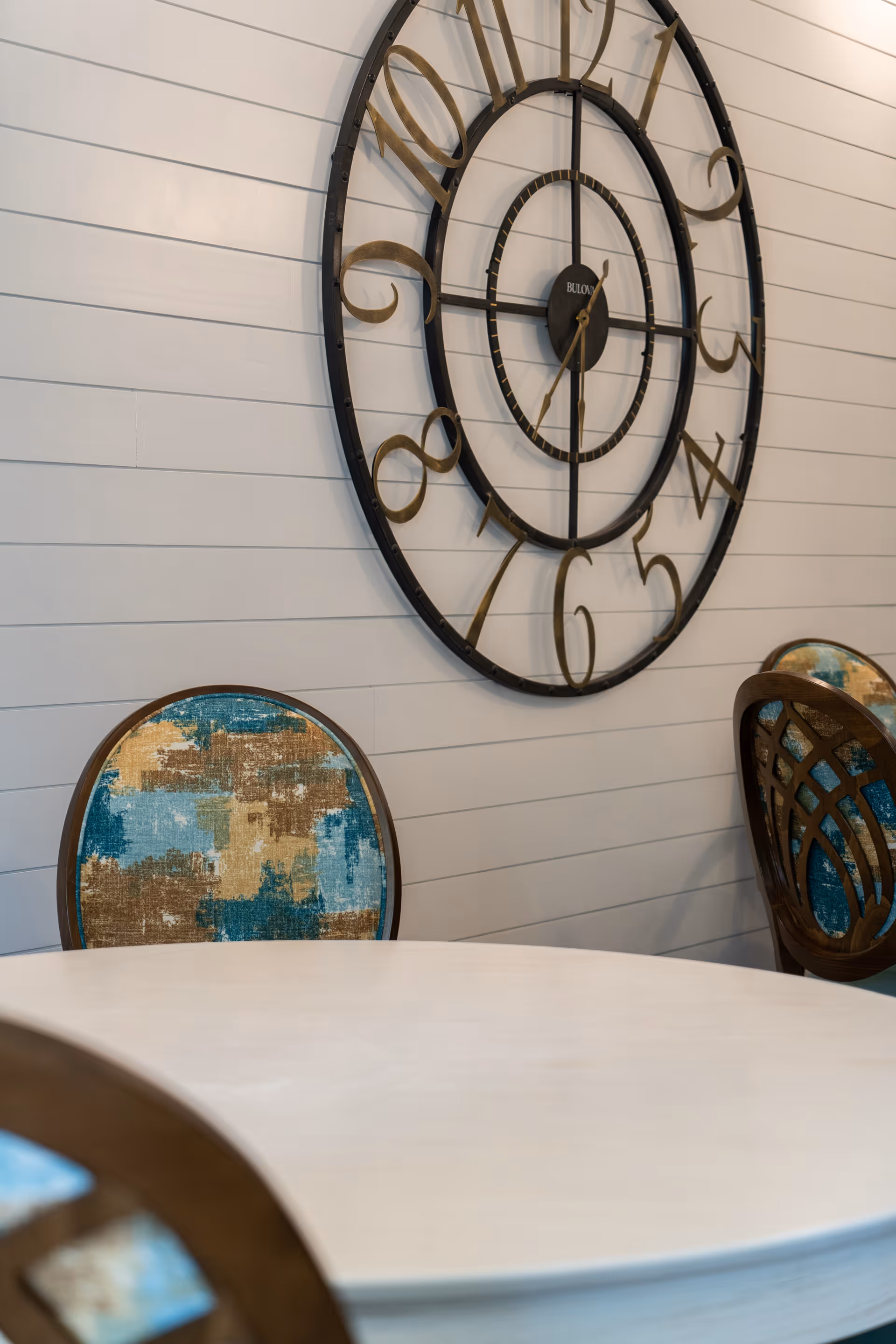 Round dining table with upholstered chairs and a large metal wall clock on a white shiplap wall.