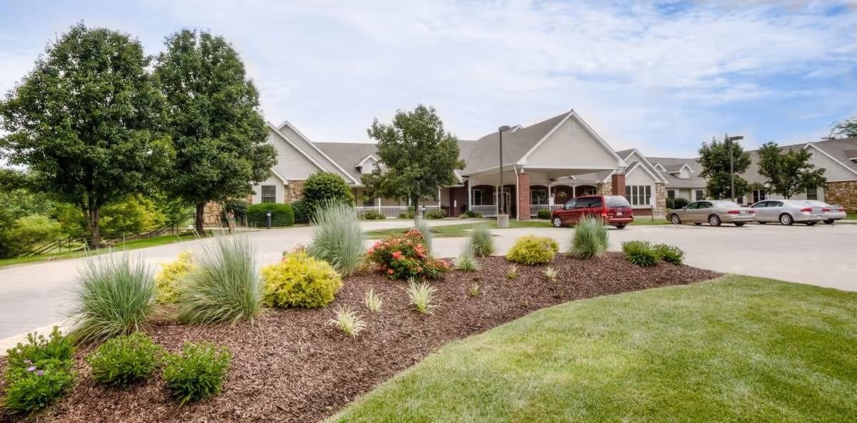 Exterior view of a senior living facility with a landscaped garden in the foreground, several trees, a parking area with cars, and a building with a covered entrance under a partly cloudy sky.