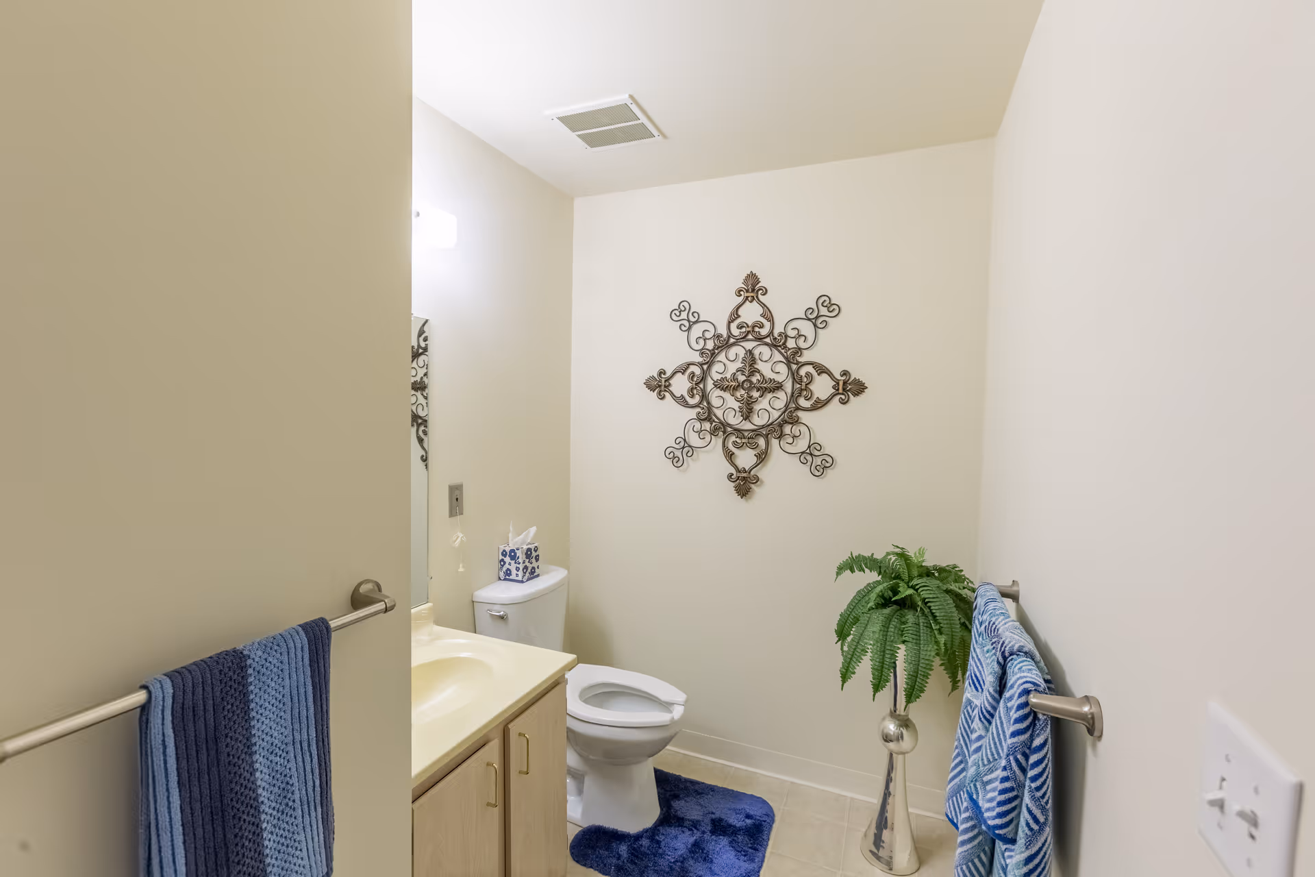 A small bathroom with a white toilet, a beige countertop with a sink, and a mirror. There are blue towels hanging on towel racks on both sides of the room. A decorative metal wall art piece is mounted on the wall above the toilet. A green potted plant in a silver vase is placed on the floor next to the toilet. The walls are painted light beige and the floor is tiled.