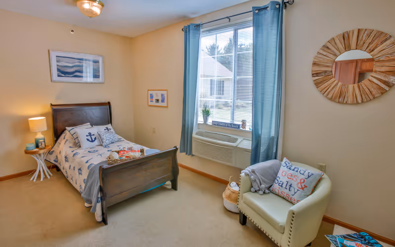 A bright furnished bedroom with a wooden single bed, nightstand lamp, armchair, and a window with blue curtains and an air conditioner.