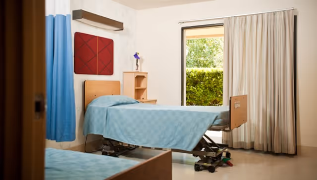 A bright patient bedroom featuring an adjustable hospital bed with blue bedding, a small bedside shelf, curtains, and a window showing hedges outside.