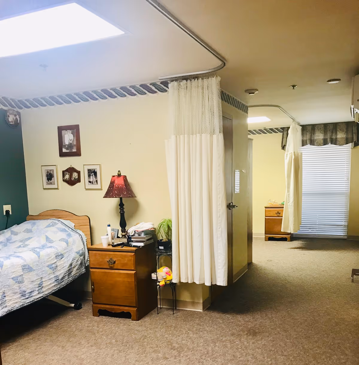 A senior living facility bedroom with a single bed covered in a blue and white quilt. Next to the bed is a wooden nightstand with a red lamp, books, and personal items. The room has beige walls with framed photos hanging and a privacy curtain partially drawn around the bed area. In the background, there is another nightstand near a window with blinds and a curtain.