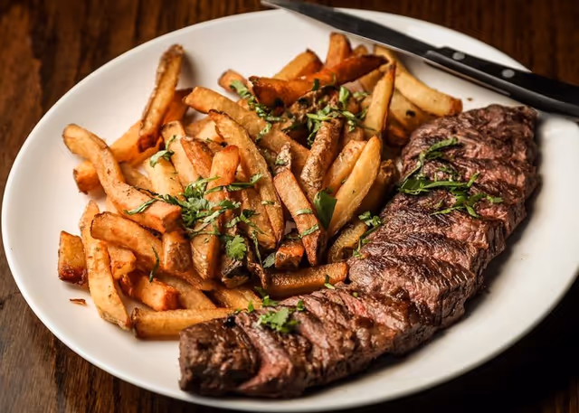 A white plate with a serving of grilled steak garnished with chopped herbs alongside a generous portion of golden-brown French fries also sprinkled with herbs, placed on a wooden table with a knife resting on the plate's edge.
