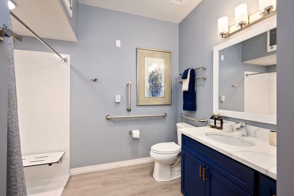 A modern bathroom with light blue walls, a white toilet, a blue vanity with a white countertop and sink, a large mirror with two wall-mounted lights above it, a shower area with a gray curtain and a foldable shower seat, grab bars on the walls, a towel rack with blue and white towels, and a framed artwork hanging on the wall.