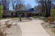 Exterior view of a single-story building with a blue awning over the entrance. A concrete pathway leads to the entrance, surrounded by landscaped areas with rocks and small plants. Leafless trees are visible in the background under a clear sky.