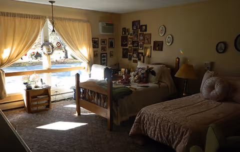 Sunlit bedroom with two twin beds, a window with curtains, and walls covered in framed photos and decorations.