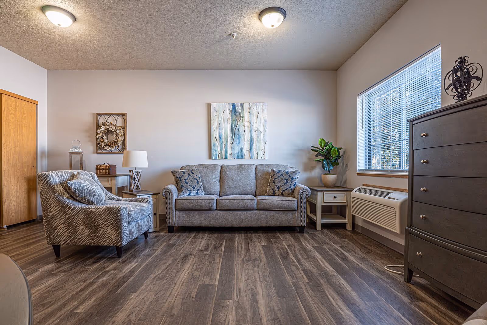 A cozy living room with a gray sofa adorned with two patterned pillows, a matching armchair with a pillow, two side tables with a lamp and decorative items, a large window with blinds, a tall dark dresser, and a painting of birch trees on the wall.