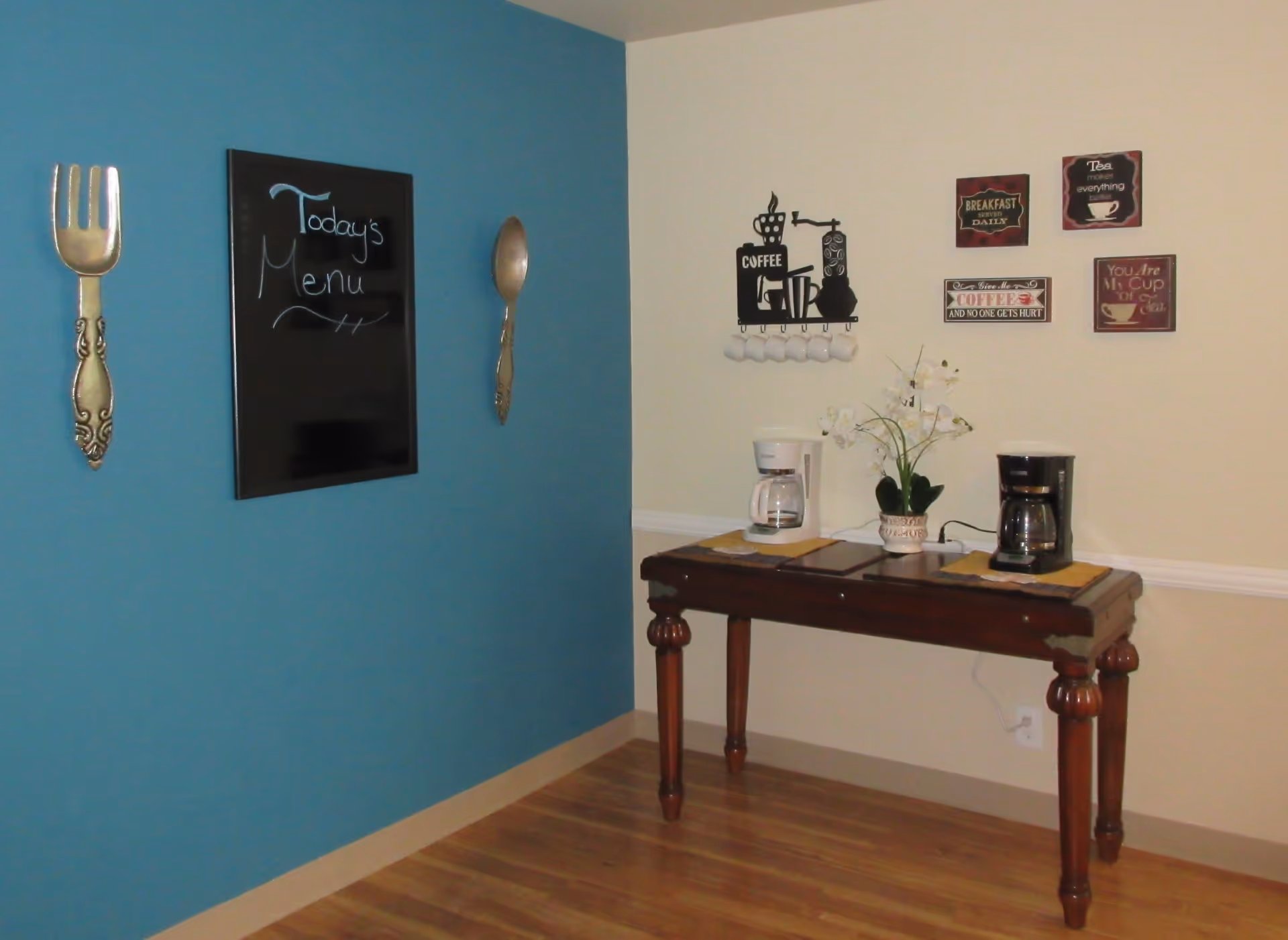 A corner of a room with a blue and beige wall. On the blue wall, there is a blackboard with the words 'Today's Menu' written on it, flanked by large decorative fork and spoon wall hangings. On the beige wall, there are several coffee-themed decorations and a wooden table holding two coffee makers and a potted plant.