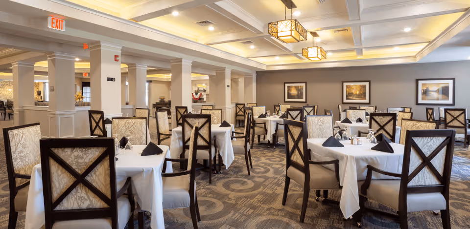 A spacious dining room with multiple round tables covered with white tablecloths, each set with black folded napkins and glassware. The chairs have dark wooden frames with patterned upholstery. The room features a coffered ceiling with decorative hanging light fixtures, neutral-colored walls adorned with framed landscape artwork, and carpeted flooring with a swirling pattern.
