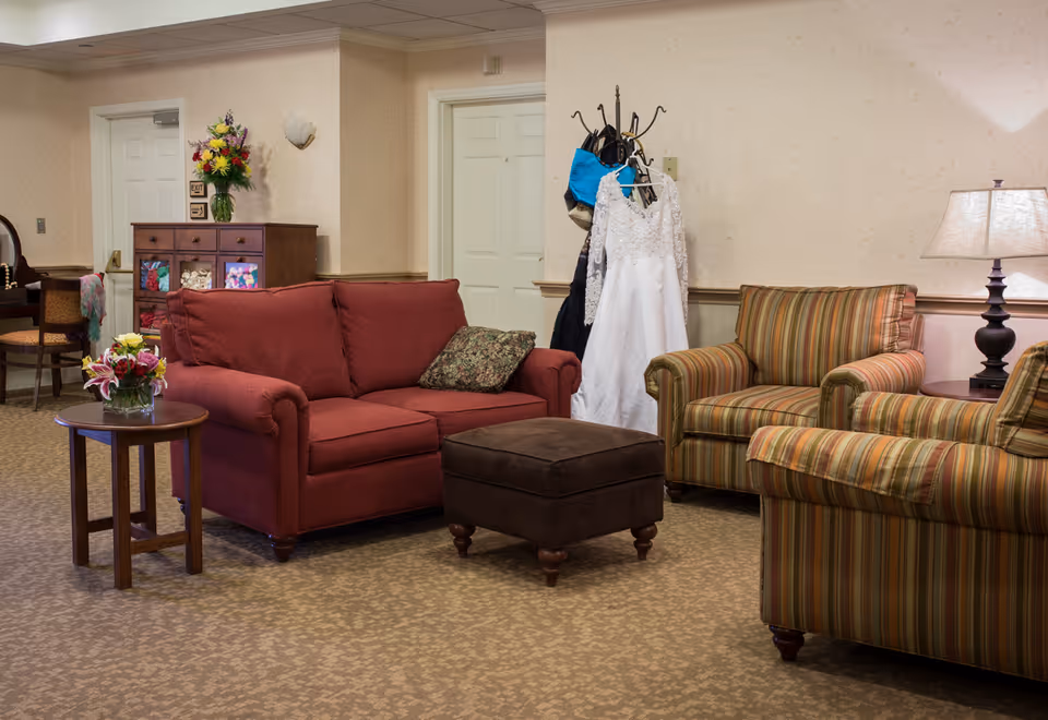 Cozy common room with a red sofa, striped armchairs, ottoman, side tables with flowers and a lamp, and a coat rack holding a white dress.