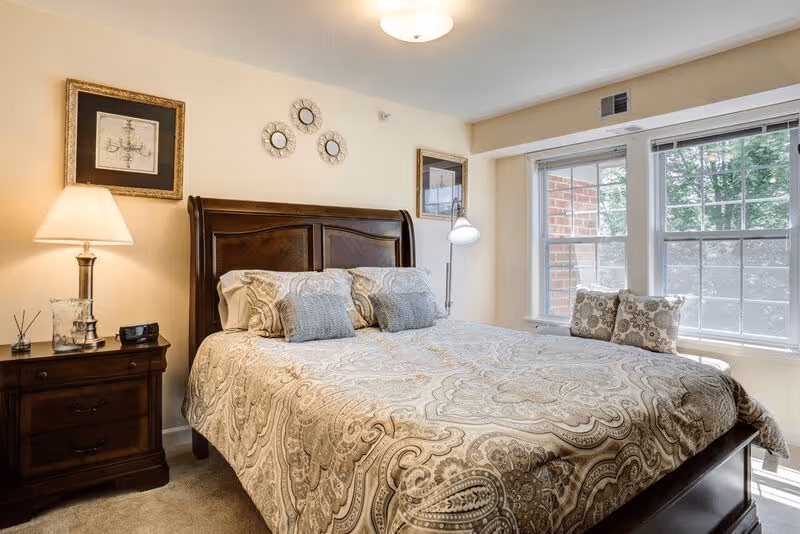 A cozy bedroom with a large wooden bed featuring a patterned beige and gray bedspread and multiple pillows. There is a wooden nightstand with a lamp, clock, and decorative items on the left side of the bed. The room has two framed pictures on the wall and three decorative round mirrors above the headboard. Large windows on the right side let in natural light and show some greenery outside.