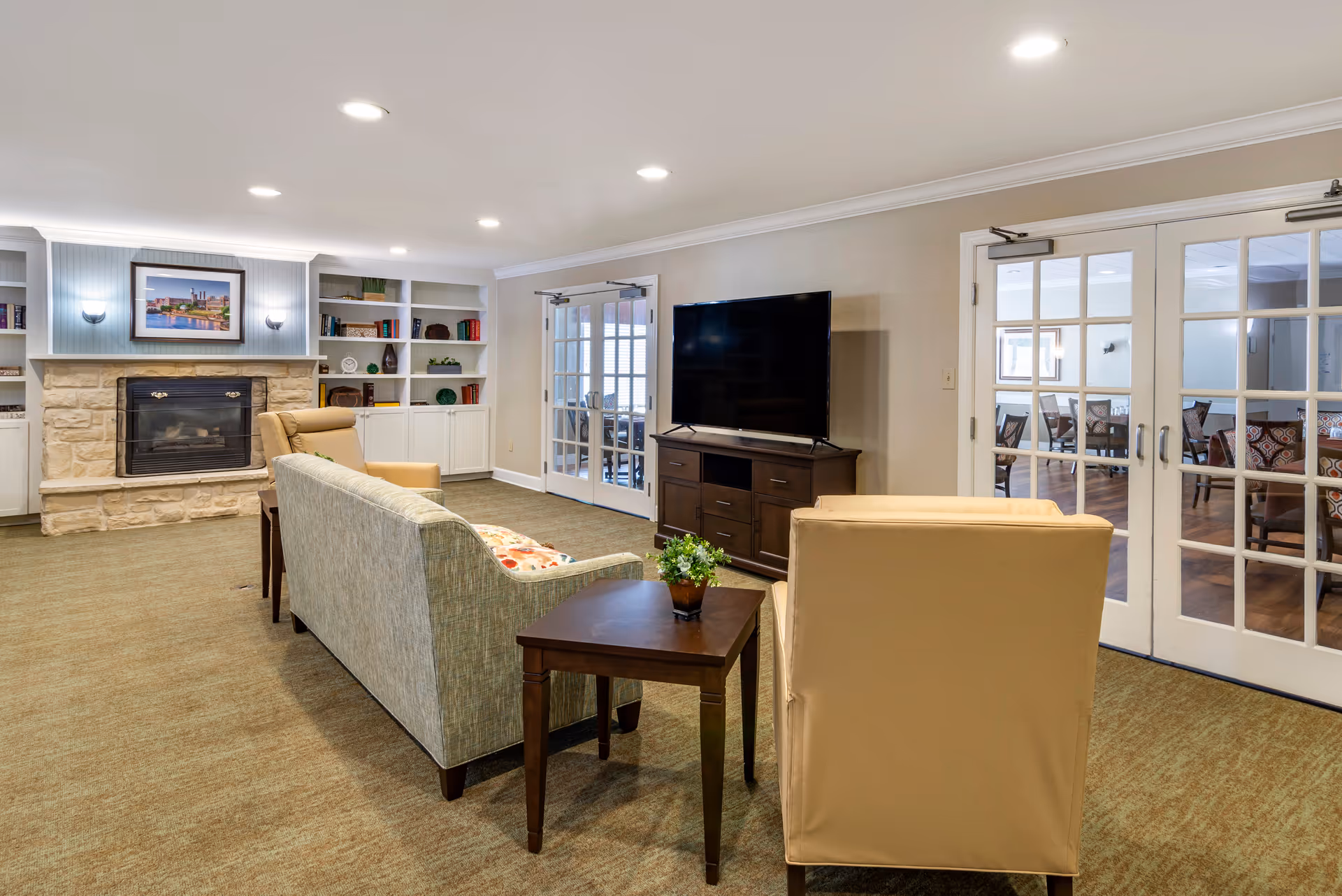 A cozy living room area in a senior living facility featuring a stone fireplace with a framed picture above it, built-in white shelves with books and decor, a flat-screen TV on a wooden stand, a beige armchair, a light green sofa, and a small wooden side table with a potted plant. French doors lead to an adjacent room with tables and chairs.