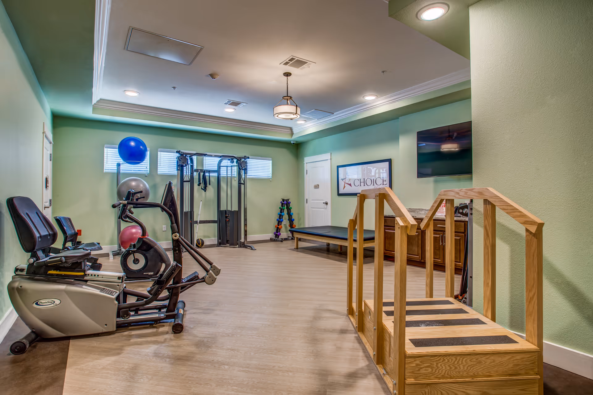 Interior view of a senior living facility exercise room with light green walls, wooden flooring, and ceiling lights. The room contains exercise equipment including a recumbent bike, resistance training machine, exercise balls, a rack of dumbbells, a wooden rehabilitation step platform with handrails, a bench, a wall-mounted TV, and a framed sign that reads 'Choice'.