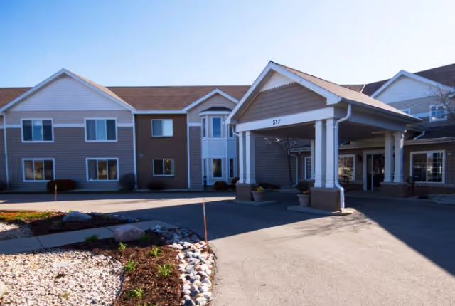Exterior view of a senior living facility named LakeHouse Fond du Lac with a covered entrance driveway, beige and white siding, multiple windows, and a landscaped area with rocks and plants in front.