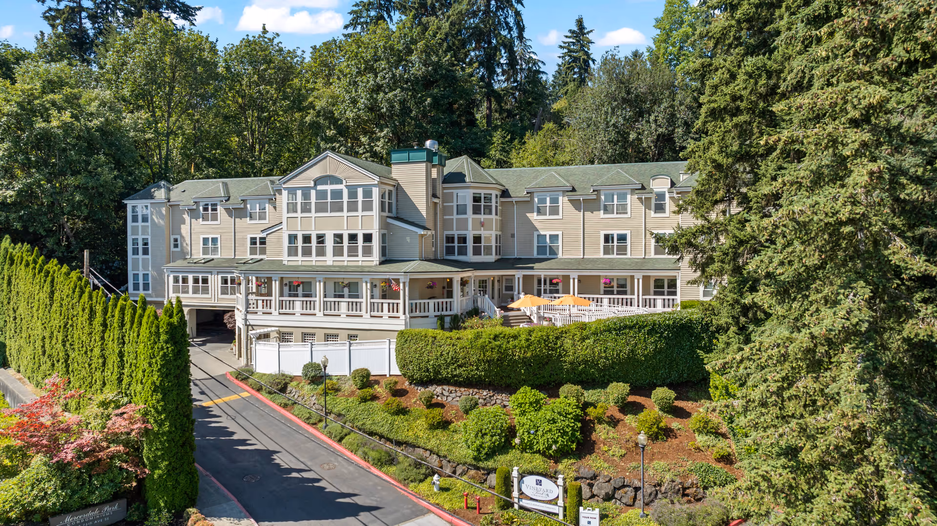 Exterior view of a multi-story senior living facility surrounded by lush greenery and trees under a blue sky with some clouds. The building has beige siding, multiple windows, and a covered porch area with outdoor seating and umbrellas. A driveway leads up to the entrance, bordered by well-maintained bushes and a sign that reads Vineyard Park.