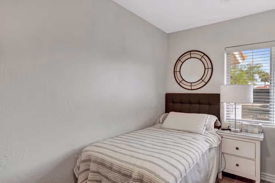 A small bedroom with a single bed featuring a striped blanket and a white pillow. Next to the bed is a white nightstand with two drawers, a modern table lamp, and a box of tissues. A round decorative mirror hangs on the wall above the bed's dark upholstered headboard. A window with white blinds lets in natural light and shows a glimpse of trees and a building outside.