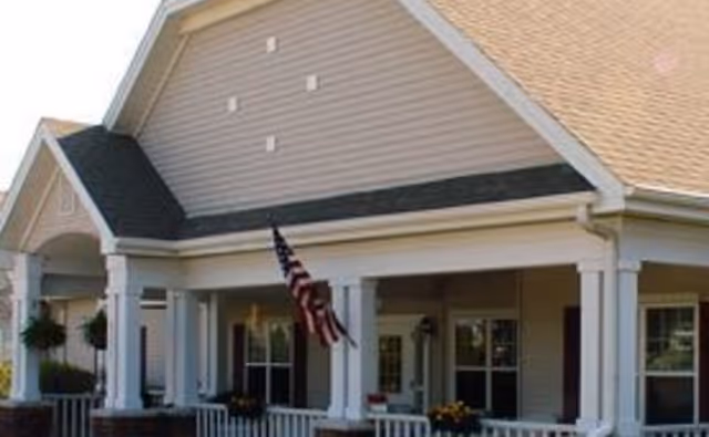 Front exterior view of a single-story building with a porch, white columns, and an American flag hanging near the entrance. The building has beige siding and a tan shingled roof.