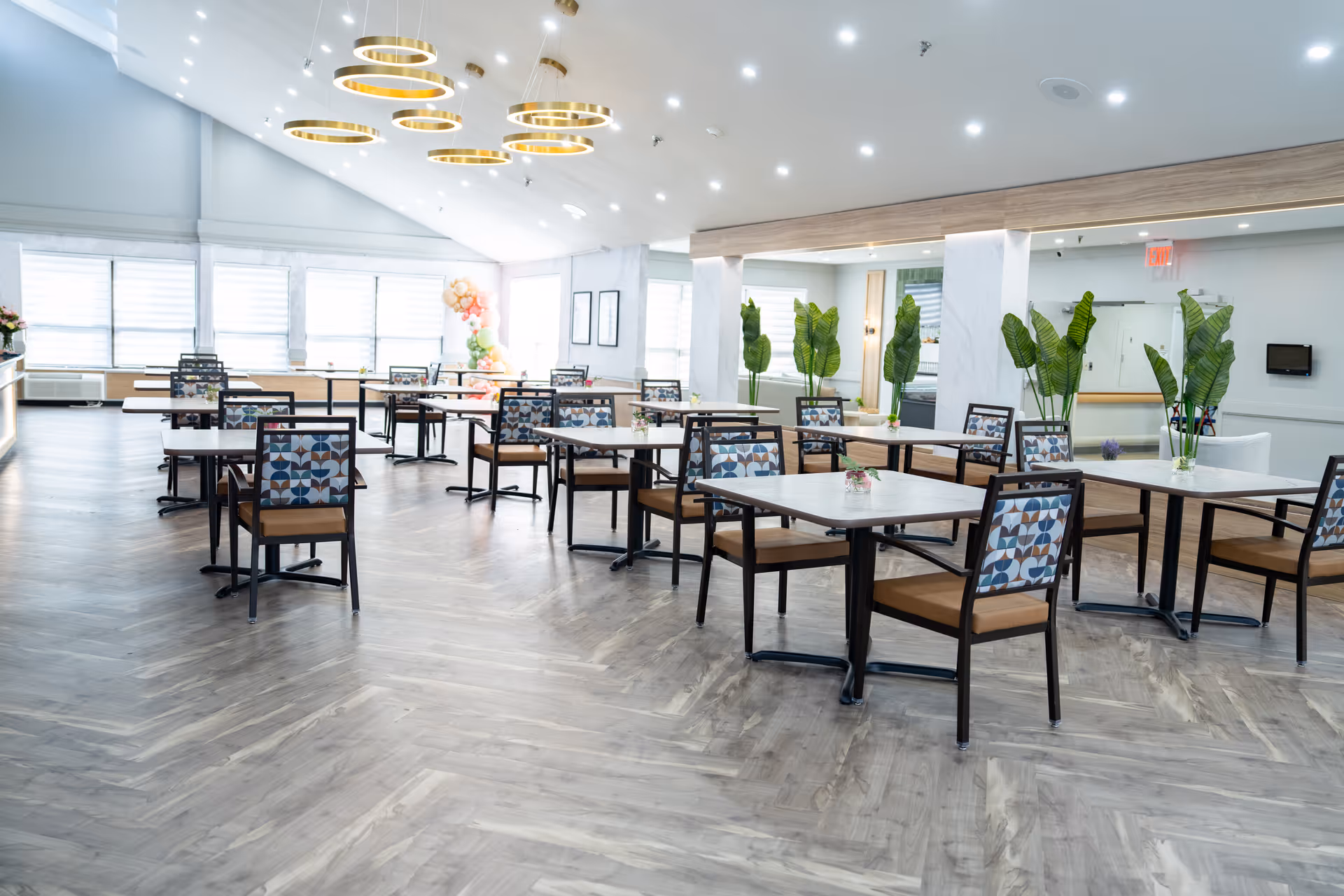 Bright, modern dining room with multiple square tables and patterned chairs, decorative plants, and circular ceiling lights.