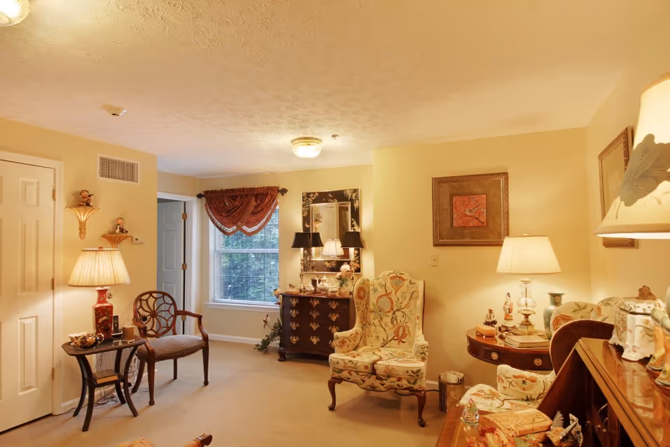 A cozy living room with beige walls and carpet, featuring a floral upholstered armchair and matching loveseat, a wooden side table with a lamp, a dark wooden dresser with a mirror and two lamps, a window with a red valance, and various decorative items including framed artwork and figurines.