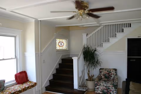 Bright interior with staircase, ceiling fan, a tall plant, and a floral chair by a window.