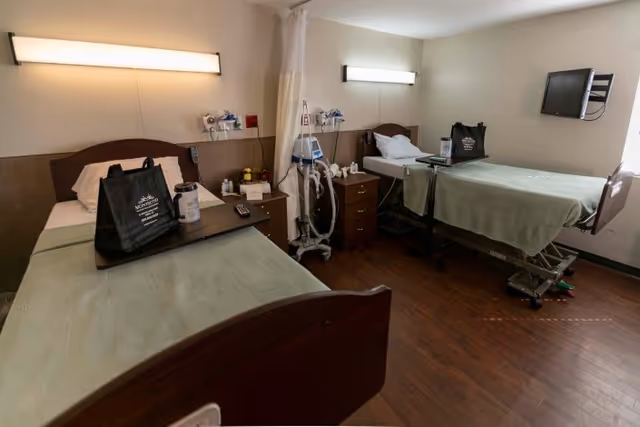 A hospital-style room with two beds separated by a curtain. Each bed has a green blanket and a black bag labeled Montecito Post Acute Care And Rehabilitation. There are bedside tables with medical equipment and supplies, a wall-mounted TV, and overhead lights above each bed.
