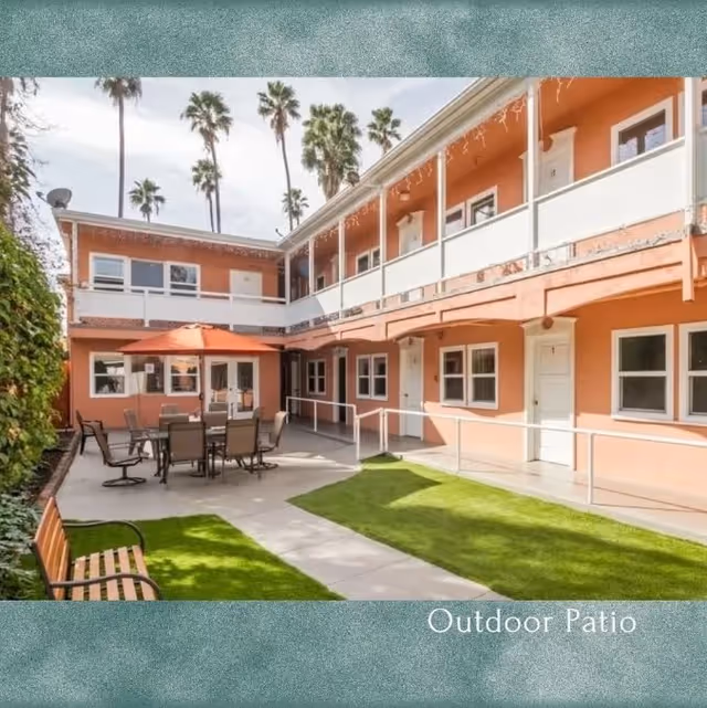 Sunny two-story courtyard patio with a dining table and umbrella, benches, grassy paths, and balconies of a peach-colored building.