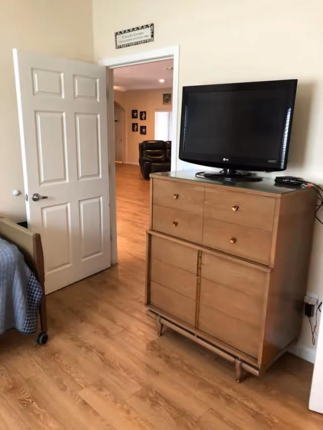 A bedroom with wooden flooring, a wooden dresser with a flat-screen TV on top, an open door leading to a living area with a leather chair, and part of a bed with a blue blanket visible on the left side.