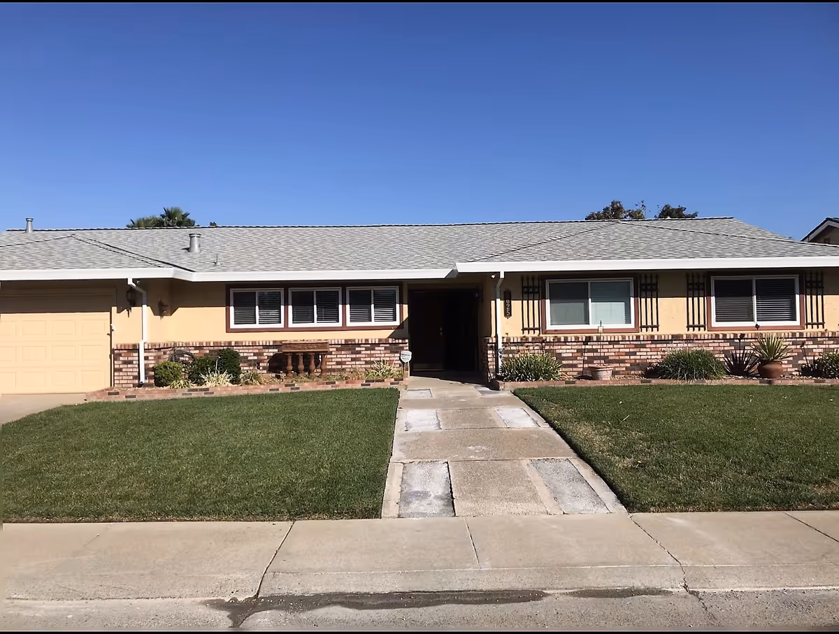 Single-story residential building with a beige exterior, brick accents, a gray shingled roof, and a well-maintained front lawn with a concrete walkway leading to the entrance.