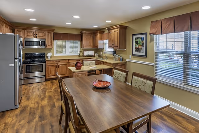 A spacious kitchen and dining area with wooden cabinets, stainless steel appliances including a refrigerator, oven, and microwave. There is a wooden dining table with six chairs, a decorative bowl on the table, and windows with brown valances letting in natural light.