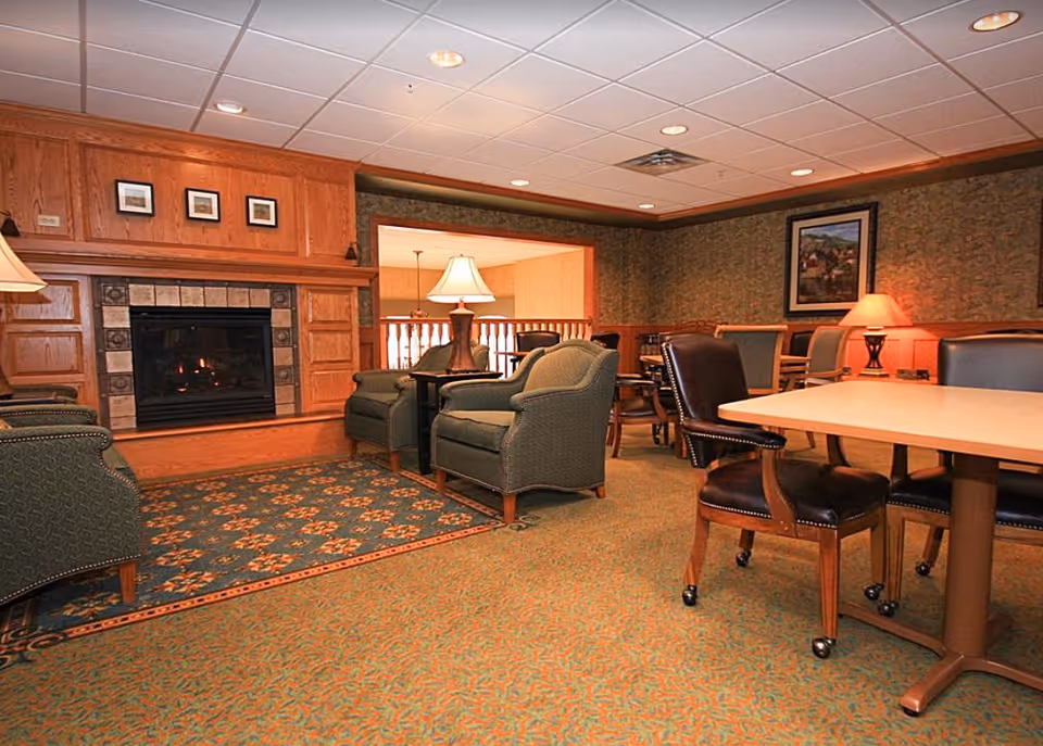 A cozy common area with a fireplace surrounded by wooden paneling. The room features several upholstered armchairs and a table with leather chairs on wheels. There are two table lamps providing warm lighting, framed artwork on the walls, and a patterned carpet covering the floor.