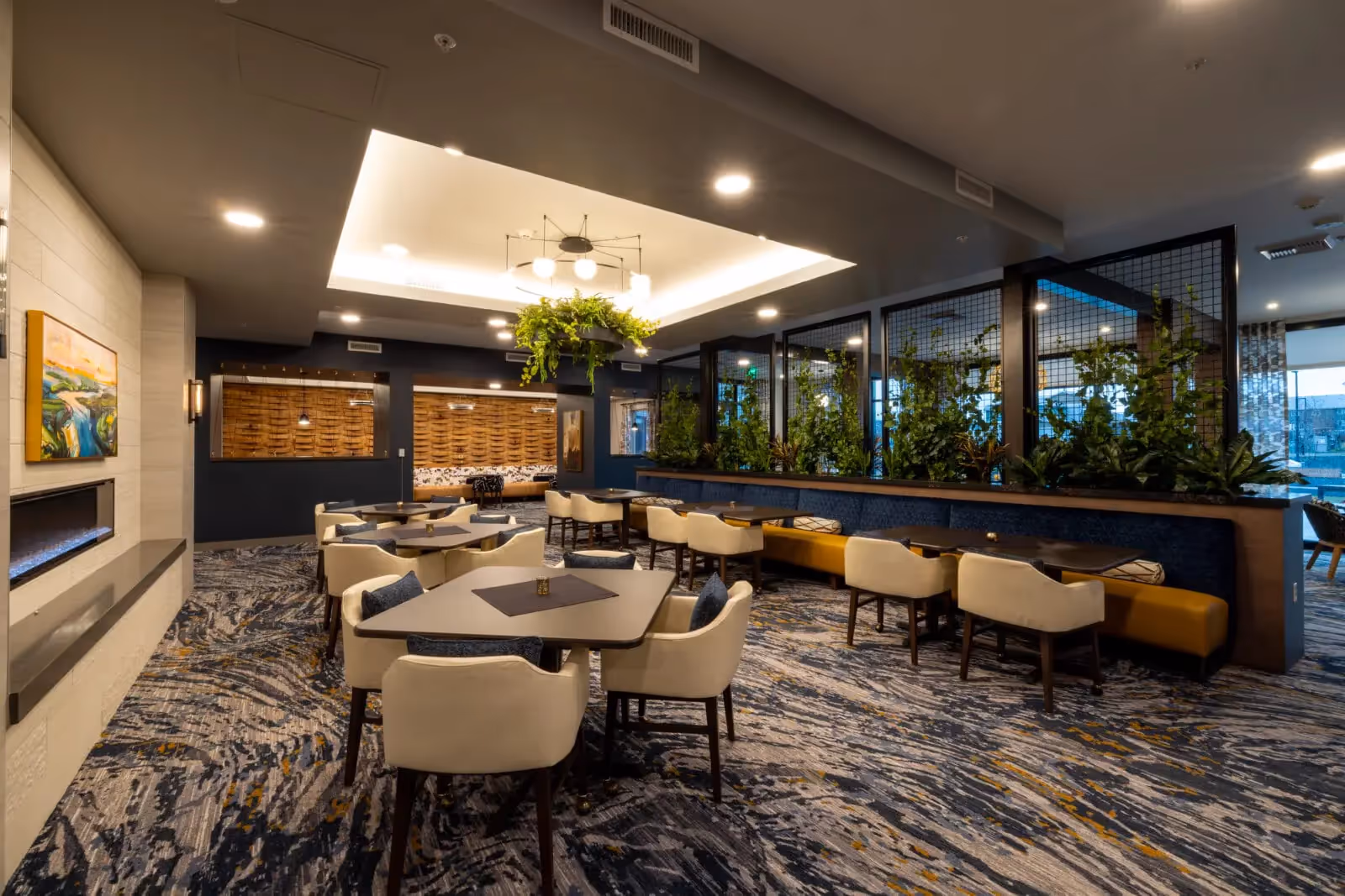 Modern communal dining/lounge area with tables, upholstered chairs, planter divider, and contemporary lighting.