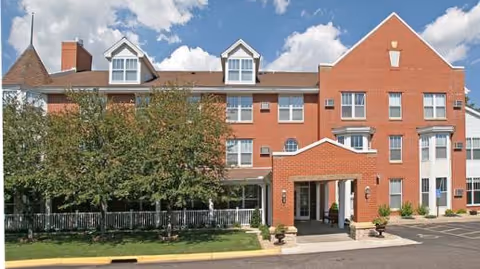 Red brick multi-story senior living building with a covered entrance, white-trim windows and landscaped front under a blue sky.