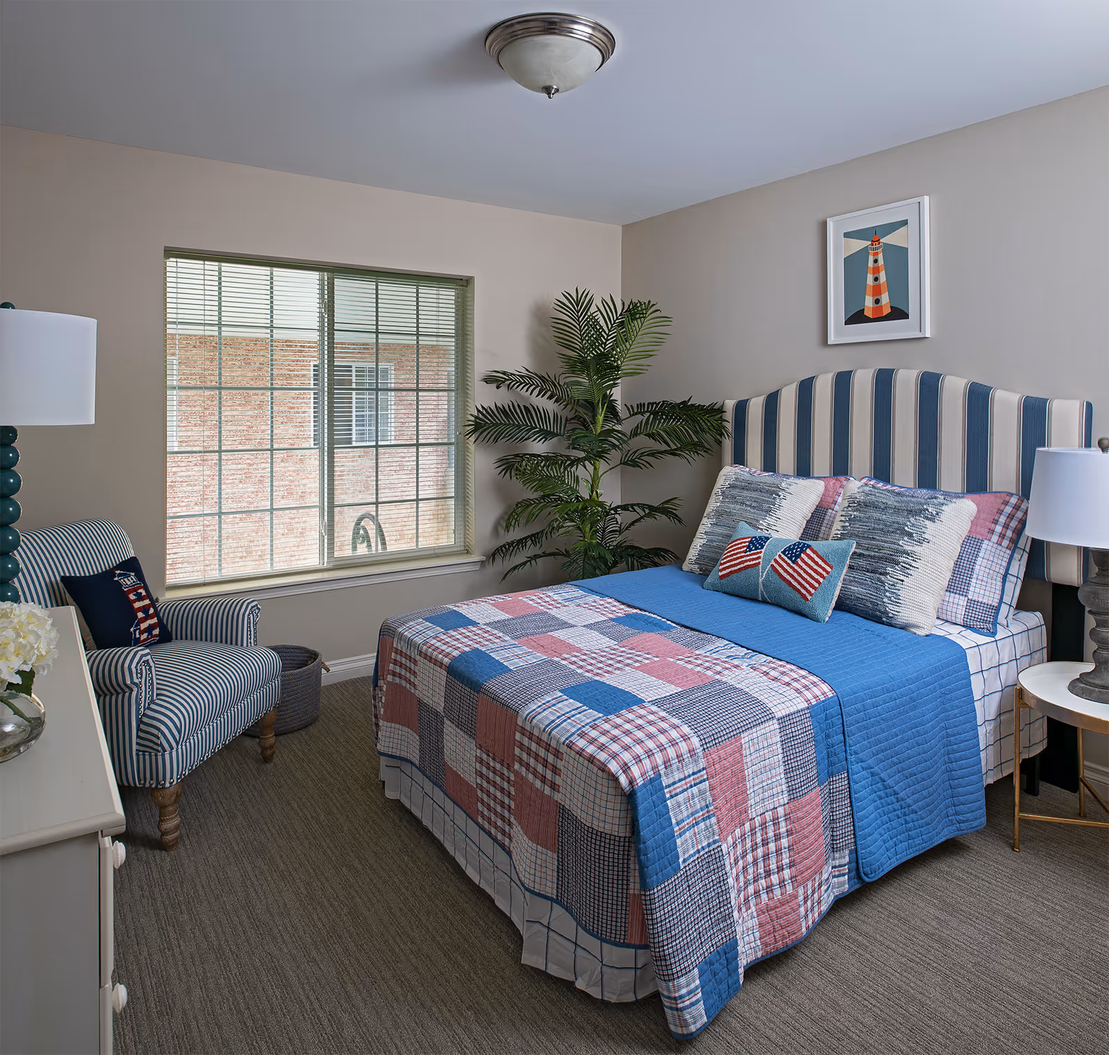 A cozy bedroom with a striped upholstered headboard and a bed dressed in a patchwork quilt with blue, red, and white colors. There are several pillows on the bed, including one with American flag designs. To the left of the bed is a striped armchair with a pillow, and a large window with blinds. A green potted plant is placed in the corner near the bed. The room has beige walls, a ceiling light fixture, and a framed picture of a lighthouse on the wall above the bed.