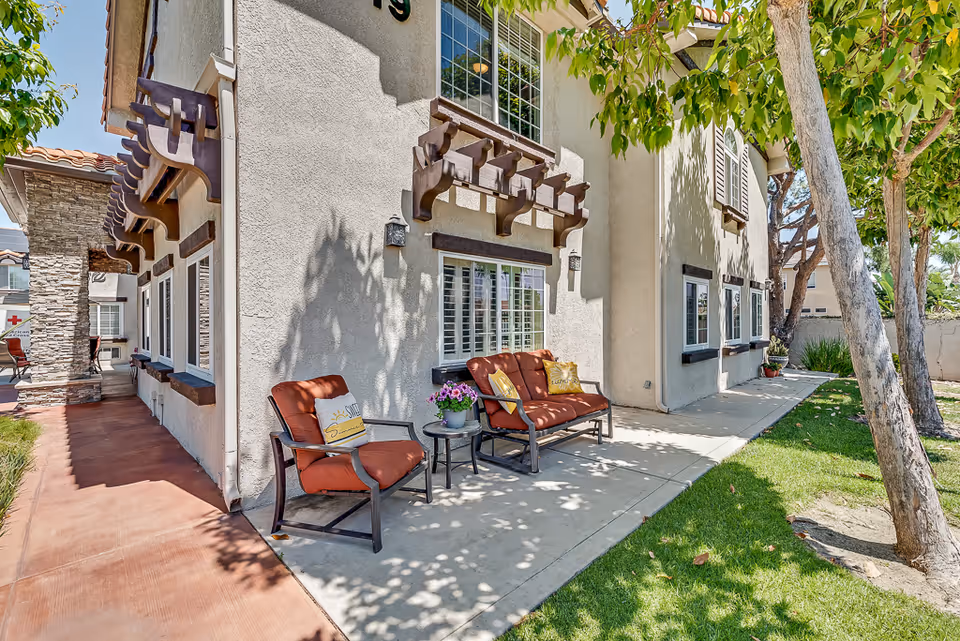 Outdoor seating area with cushioned chairs and a small table and potted flowers along the side of a two-story stucco building under trees.