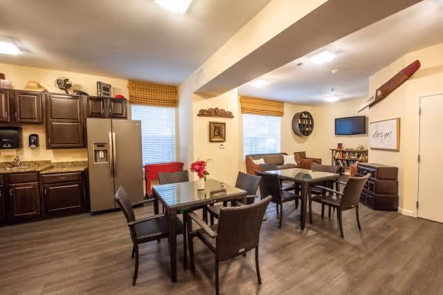 Interior view of a senior living facility common area featuring a kitchen with dark wooden cabinets, a stainless steel refrigerator, and a granite countertop on the left. In the center and right, there are two glass-top dining tables with wicker chairs around them. The background shows a cozy living area with a sofa, a wall-mounted TV, a bookshelf, and decorative wall art including a wooden propeller and framed pictures. The room has wood flooring and large windows with woven blinds.