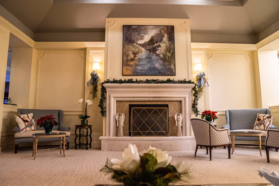 A cozy living room area with a central fireplace decorated with greenery and two angel statues on the mantel. Above the fireplace hangs a painting of a river scene. On either side of the fireplace are wall sconces with soft lighting. The room features two blue upholstered armchairs with bird-patterned pillows, two small tables with red poinsettia plants, and additional seating with patterned chairs. The carpet is light-colored with a subtle pattern, and the walls are painted in warm neutral tones.