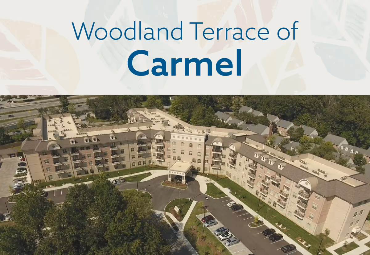 Aerial view of Woodland Terrace of Carmel, a multi-story senior living facility surrounded by trees and nearby residential houses, with a parking lot and driveway in front.