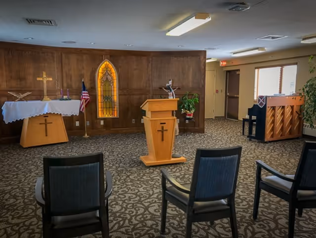 Interior of a small chapel or worship room with wooden walls, a podium with a cross, an altar with a cross and candles, an American flag, a stained glass window, several chairs arranged facing the podium, and a piano near a window.