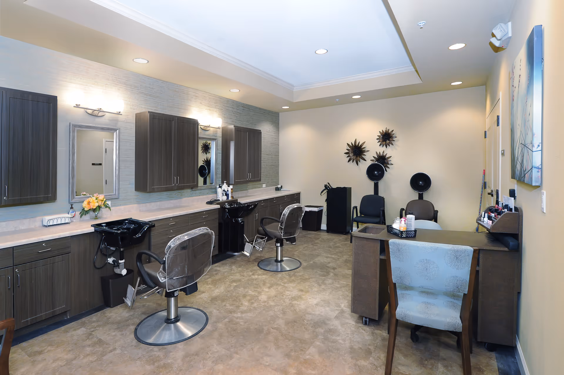 Interior view of a senior living facility's hair salon with two styling chairs in front of sinks and mirrors, dark wood cabinetry, a desk with a chair, and hair drying stations against the far wall.